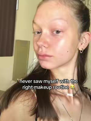 The cover image shows a close-up of a young woman's face, angled slightly to the side. She has light skin, with some redness visible on her cheeks, presumably before makeup. Her dark hair is pulled back, and she is wearing small hoop earrings. The background is a simple interior setting, with what appears to be a dark curtain and a cabinet, the lighting is soft and natural. A text overlay at the bottom of the image reads, "never saw myself with the right makeup routine".