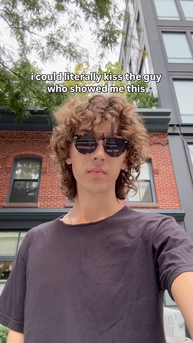 A young man with curly brown hair and sunglasses stands in front of a brick building and a tall modern building. The text overlay reads, 'i could literally kiss the guy who showed me this'. He wears a dark t-shirt and a silver chain.