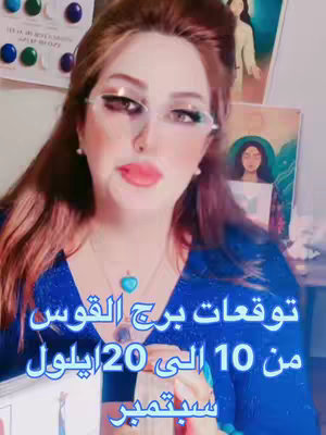 A woman with long brown hair and wearing clear glasses is the main subject in the image. She is wearing a blue top and has a necklace with a teal pendant. The background appears to be a room with a whiteboard or similar item, and a painting with an image of a person.