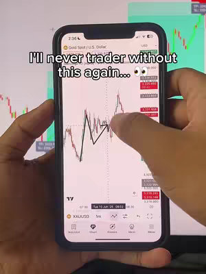 A person is holding a mobile phone with a trading application open. The phone screen displays a graph with lines and data points in front of another screen, likely a computer monitor. The person's hands are visible, holding the phone and interacting with the screen.