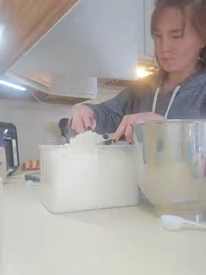 A person is shown in a kitchen setting, scooping a white substance, likely flour or sugar, from a rectangular container with a black scoop. They wear a gray long-sleeved shirt. A large metallic mixing bowl and a stand mixer are partially visible on the countertop.