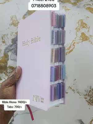 A person's hand holds a light pink Bible with multiple colored tabs along the right side. Text and numbers are overlaid. The background has a marbled effect.