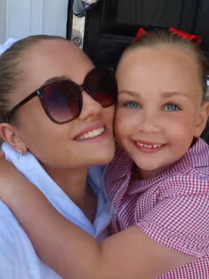 A close-up photo of two females embracing and smiling. The woman on the left is wearing sunglasses. The small girl on right is hugging her. 