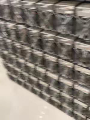 The image shows a wall of stacked ASIC miners, devices used for cryptocurrency mining. They are arranged in rows and columns, filling the frame. The setting appears to be an industrial or warehouse environment, with a plain, light-colored floor providing contrast to the metallic gray devices.
