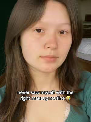 A selfie-style shot featuring a young woman with long brown hair and a fair complexion. She is centered in the frame, looking directly at the camera with a neutral expression. Overlayed text at the bottom reads, 'never saw myself with the right makeup routine.' The background is a soft, out-of-focus blend of green and tan tones, with hints of a white bed on the upper right.