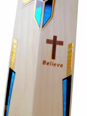The image shows a close-up of a cricket bat angled diagonally. The bat is primarily off-white with colorful accents. Along its edges, there are gold and blue stripes and shapes. In the center, there is a brown cross and the word "Believe" in a similar brown font. The overall composition is striking with its religious symbolism within sporting equipment.