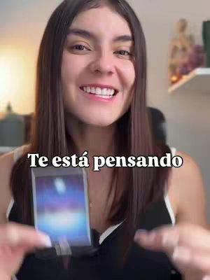 A young woman with long, straight brown hair and a white top smiles directly at the camera, holding a smartphone that displays a tarot card. The text "Te está pensando" is overlaid on the image in large, bold, white letters. The background is softly blurred, suggesting an indoor setting, and the overall composition is brightly lit, emphasizing the woman's engaging expression and the central focus of the tarot card.