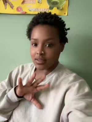 A woman with short, curly black hair looks directly at the camera, wearing a beige sweatshirt. She holds her hand up near her face with her fingers spread. The background is a light green wall, with a yellow poster and a green cartoon character sticker in the top of the frame.