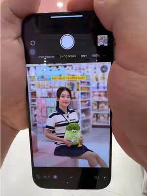 A person is holding up a phone displaying an image of a well-lit woman in what appears to be a retail store. The phone is in the foreground, being held in both hands. The phone screen shows a woman holding a plush toy while a store is in the blurred background. The phone's interface is partially visible. Text in Vietnamese appears to be associated with the phone's settings, and photo-taking tools are visible.