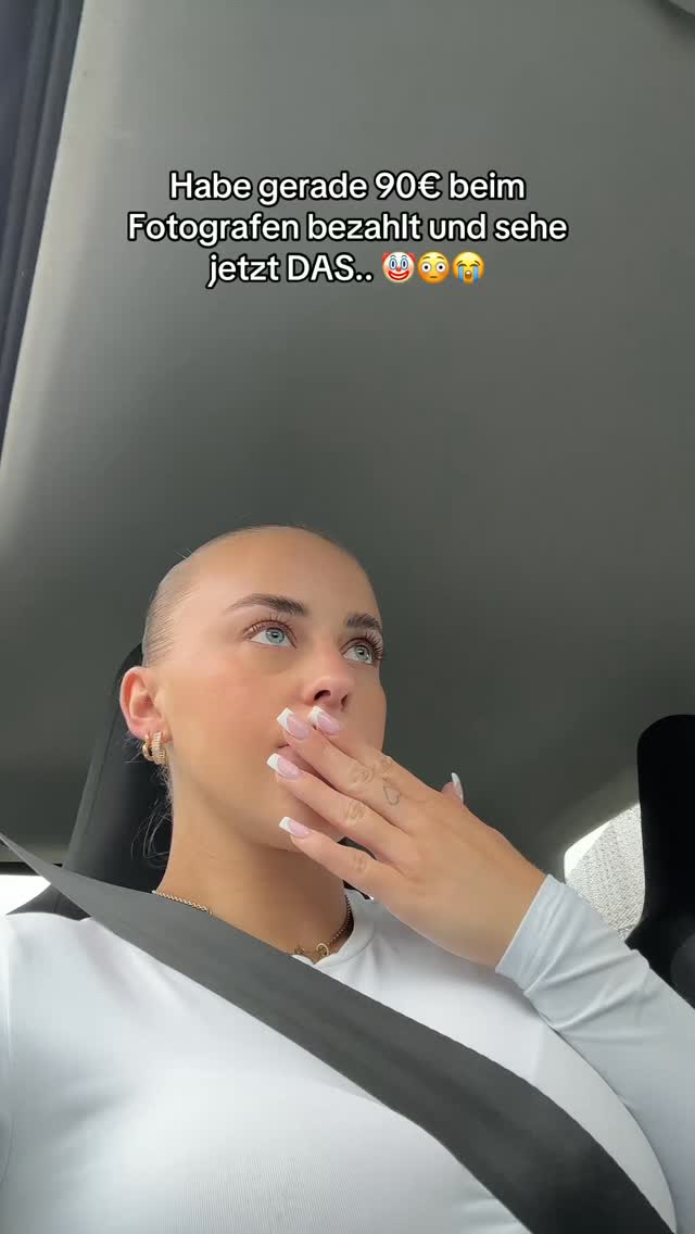 A selfie taken from inside a car. A woman with fair skin and light blue eyes is covering her mouth with her right hand, expressing surprise. Her hair is pulled back, and she wears a white top and gold earrings. The background is a blurred interior, and text is overlaid. The overall composition suggests a reaction to something.