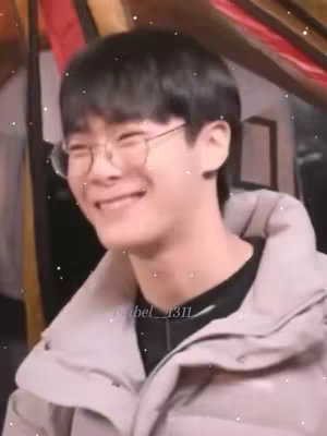 A close-up shot of a young man with black hair and glasses smiling broadly. He is wearing a light-colored jacket over a black shirt. The background is slightly blurred, with some red and yellow.