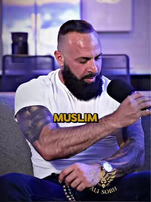 A man with a full beard and a white t-shirt is holding a microphone and wearing a watch. The word 'MUSLIM' is displayed in a yellow highlighted banner. Tattoos are visible on his arm.