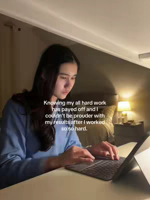 A young woman is seated at a desk, working on a laptop. She is looking down at the screen, and the background is dark. The image is warmly lit.