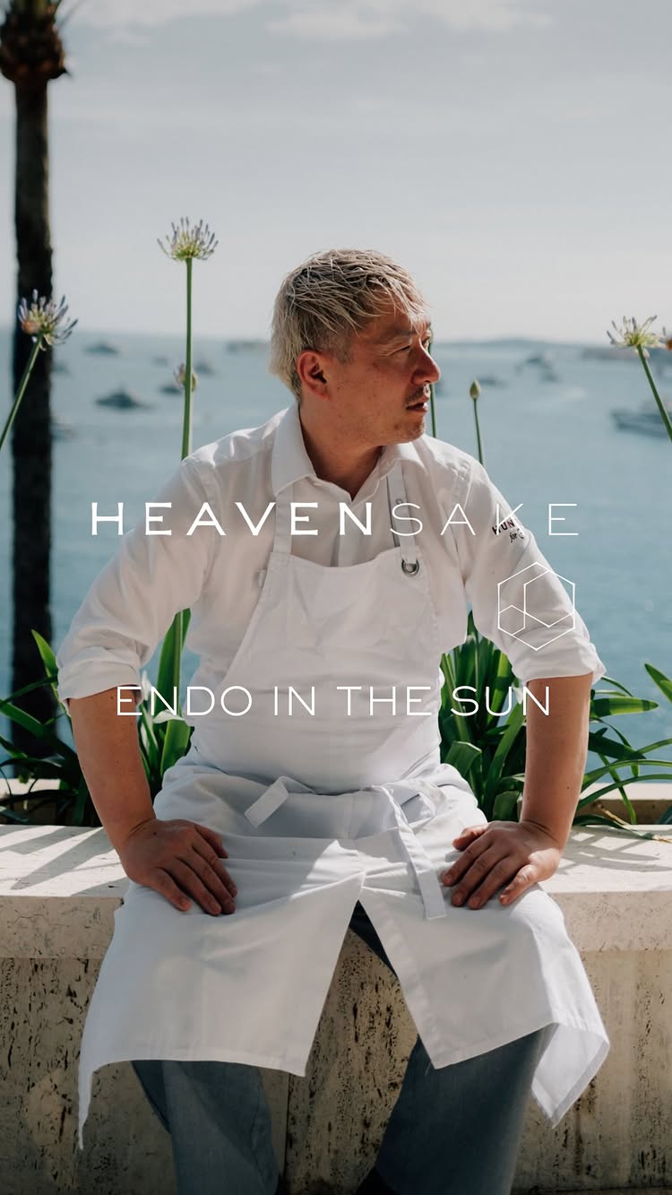 A man in a white chef's coat and apron is seated on a stone ledge overlooking a scenic background of water and distant shoreline. Text overlays the image. The composition is bright with natural light.