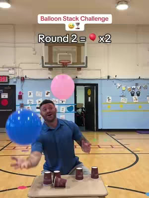 The image features a man in a blue shirt in a gymnasium setting. He appears to be interacting with a table holding cups, with a pink balloon and a blue one also visible. The background includes a basketball hoop.