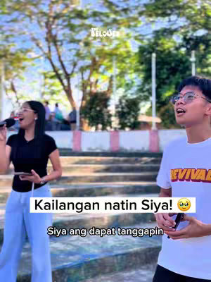 The image captures two individuals standing outdoors near stairs, with a backdrop of green trees and a clear sky. The woman, on the left, is wearing a black top and light blue pants and holding a microphone. The man on the right is wearing a white t-shirt and glasses, and looks upwards. Overlayed text in a white box emphasizes a message in the video's theme.