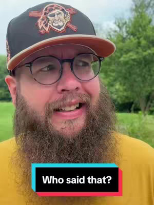A close-up shot of a man wearing glasses and a baseball cap. His beard and mustache are thick and full, and he is smiling with a slightly open mouth. Overlayed on the image at the bottom is a black and cyan rectangle with white text inside. The background consists of some green foliage.