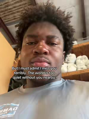 A close-up shot of a young Black man looking directly at the camera with a slightly sorrowful expression. He has tightly curled dark hair. Overlayed text is visible, and the background is somewhat blurred, suggesting an indoor setting with wooden shelving.