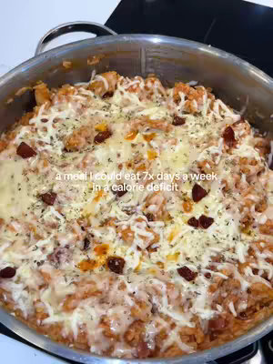 A close-up shot of a pan filled with cheesy chicken and chorizo rice. The dish features a reddish-orange rice base, mixed with visible pieces of chicken and chorizo. The top is generously covered with melted cheese and herbs. The pan sits on a black stovetop, and the overall lighting is bright and highlighting the colors of the food.