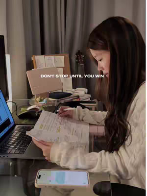 A person is studying at a desk, angled from the side. The lighting is soft, suggesting dim indoor light. There is text overlaid at the top that says "DON'T STOP UNTIL YOU WIN". The person is looking at a document with text, and there is a laptop and a phone on the desk. The scene conveys a study environment with a motivational quote.