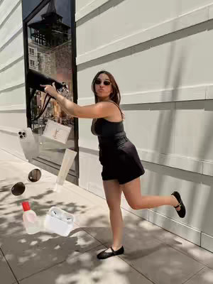 A young woman in a black outfit and sunglasses is captured mid-leap, throwing items from a black bag. The background features a modern building facade and reflections in a large glass window. Items spilling from the bag include a phone case, a designer wallet, a makeup item, a perfume bottle, AirPods, and a mirrored compact. The overall composition is bright and sunny.