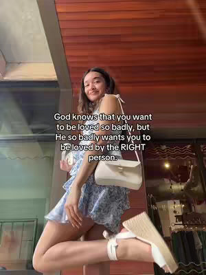 A young woman stands outdoors, leaning against a wall, smiling at the camera. She's wearing a light blue dress with small flower prints and white sandals, holding a white purse. Text is overlaid on the image, with wooden panels on the background.