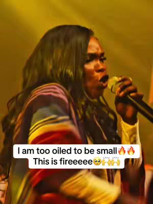 A woman with long, dark hair sings into a microphone. She is wearing a colorful shawl. Text overlayed at the bottom states: "I am too oiled to be small".