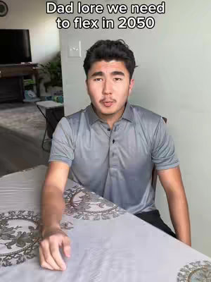 A young man in a gray polo shirt is seated at a table. Text at the top of the image says 'Dad lore we need to flex in 2050'. The man is looking directly at the camera. The background includes a light wall and a glimpse of another room. The table is covered with a patterned tablecloth.