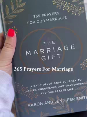 The cover of a book titled "The Marriage Gift." It is held by a person with red nail polish. The book has a dark gray cover with gold-colored accents, including the title text, a design of intertwined rings and floral elements. Additional text includes "365 Prayers for Our Marriage", and author names.