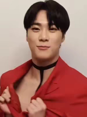 A close-up shot features a smiling man with a black choker and a red blazer, with a neutral background. He's holding open the blazer, with an expression of confidence.
