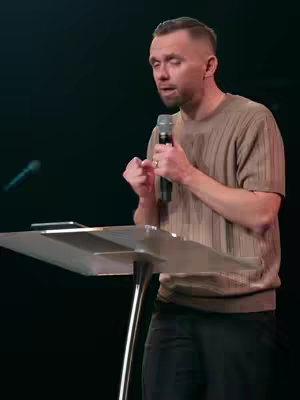 A man in a brown ribbed shirt stands behind a transparent podium, holding a microphone. The background is dark. He appears to be giving a speech or presentation, gesturing with his hands. The lighting seems to be from above.
