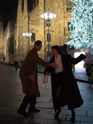A night scene in a European city square featuring two individuals in a moment of interaction. The background includes a lit-up grand building, likely a cathedral, and a large decorated Christmas tree. The foreground shows a man extending his hand, gesturing towards a woman who has one arm open. The lighting is dim with streetlights providing illumination, setting a festive atmosphere.