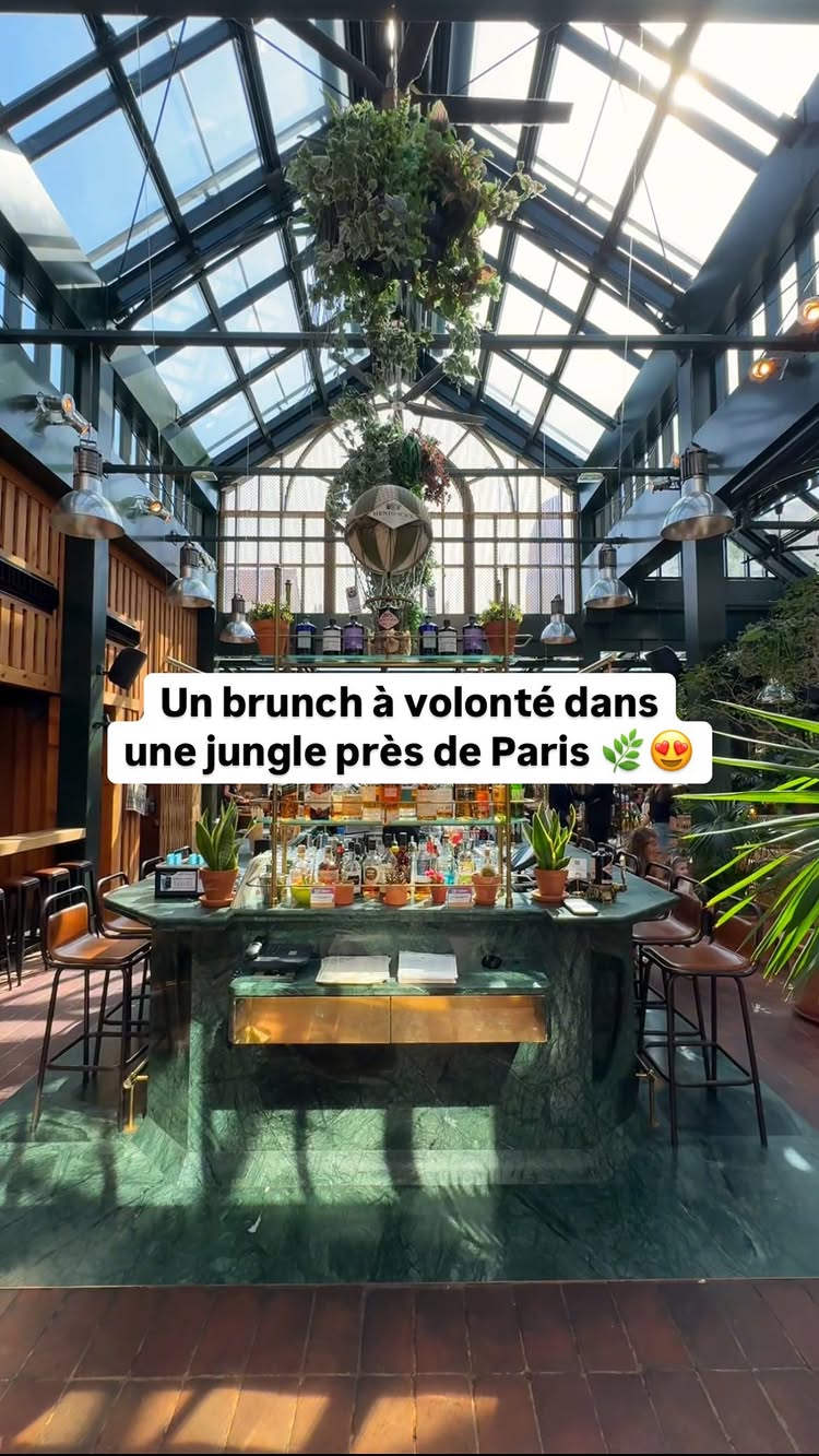 The image shows a stylish brunch restaurant interior. The focal point is a long, green marble bar filled with bottles and plants. Above, large glass skylights illuminate the space, with hanging plants adding to the jungle aesthetic. The overall composition uses a wide angle to capture the environment.