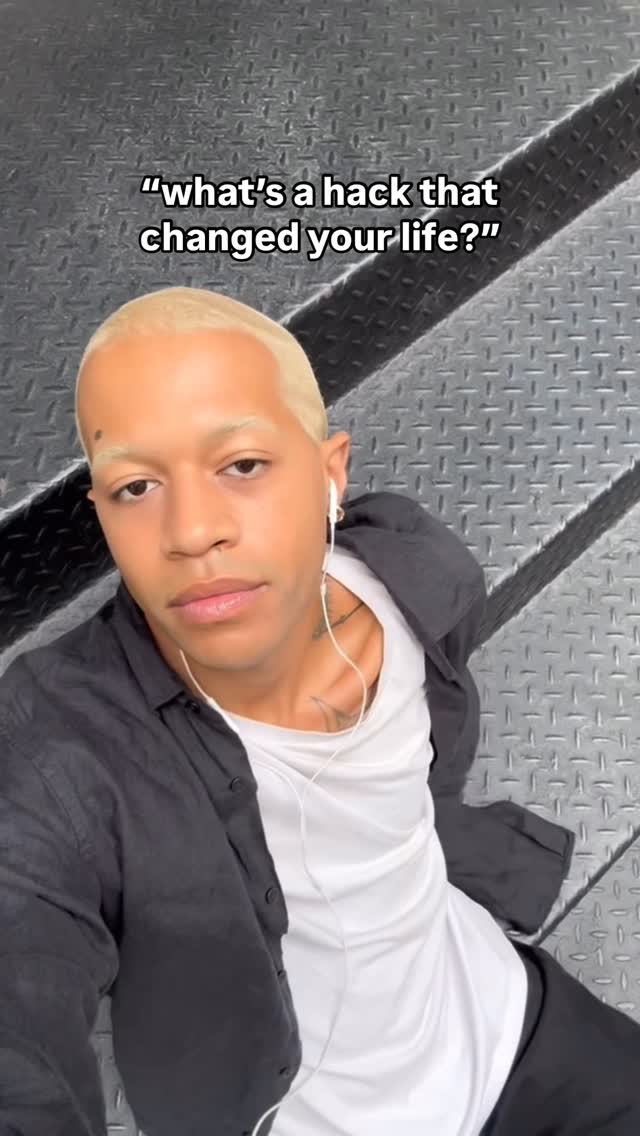 A person with blonde hair wearing a black shirt over a white t-shirt looks toward the camera. Their face takes up a large portion of the image. They have in-ear headphones in. The background consists of a gray metal pattern.