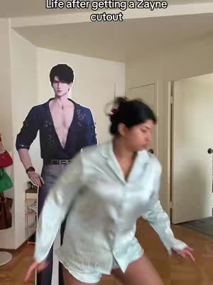 A woman dances in a room with a large cutout of a male anime character. The cutout has a dark blue shirt and grey pants. The woman is wearing a light blue pajama set and the room has wooden floors and is brightly lit.