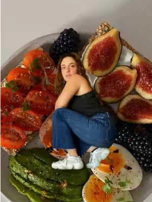 A woman sits amidst a colorful arrangement of food on a large, oval-shaped plate. The food includes halved tomatoes, sliced avocados, halved figs, blackberries, and halved hard-boiled eggs. The woman is wearing a black top, blue jeans, and white sneakers. She looks towards the camera.
