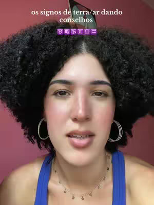A woman with dark curly hair and light skin gazes towards the camera. The background is a soft pink, and a text overlay is positioned behind her head. She wears a blue tank top, silver hoop earrings, and a delicate necklace. The composition is a close-up, emphasizing her facial features as she speaks.