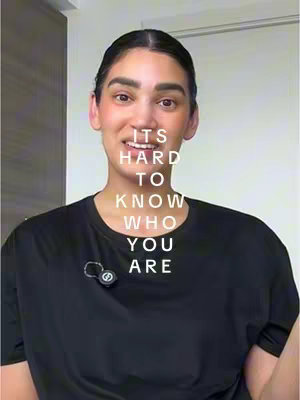 A woman with dark hair pulled back and light makeup is centered in the frame. Text overlays her face, and she wears a black shirt and a small microphone. The background is a white wall.