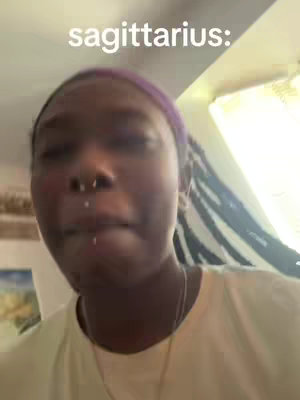 A selfie-style shot featuring a person with a purple headband and multiple facial piercings. The person is centered, with a slightly blurred background suggesting an indoor setting. The text overlay "sagittarius:" appears in the upper left corner, in a clean sans-serif font, providing context linked to zodiac signs.