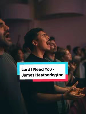 A vertical image showing a group of people, many with their eyes closed and mouths open, singing. A text overlay in a colorful bar indicates the song title and artist at the bottom. The overall lighting is soft and warm, suggesting a concert or religious setting.