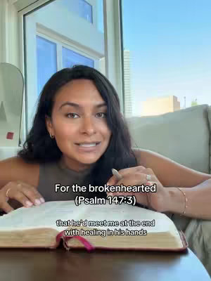 A woman is seated indoors, holding an open book with text overlaying the image. The background includes a window with bright daylight.