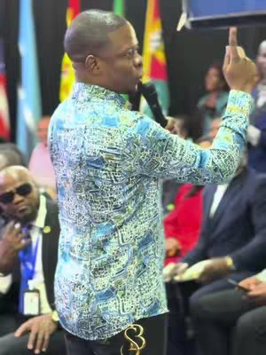 A man in a patterned shirt is speaking into a microphone in front of a crowd and various flags. He is raising his right hand.