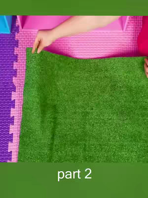 The image is taken from an overhead perspective. A green, artificial grass mat covers the majority of the frame. The bottom of the image shows a green background with the words "part 2" in white text. Two hands, belonging to what appears to be a person, are visible in the upper portion of the image reaching down to the green turf. The hands are touching the turf, seemingly holding the material flat. In the background, at the top, there's a pink surface, possibly a table or a mat, with a partial view of a blue element on the left side.