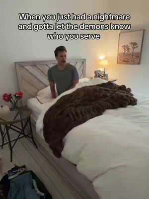 A young man in a gray shirt is standing near a bed, with a fluffy brown blanket draped over the white comforter. The room is simply decorated with light-colored walls; a framed piece of art hangs on the wall. A small table holds a vase of red flowers.