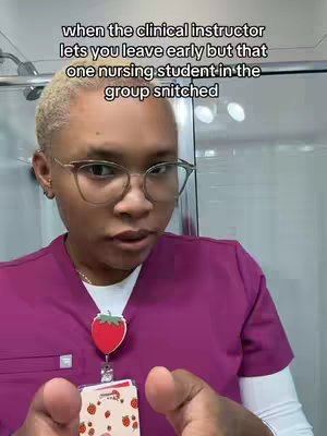 A woman with short blonde hair wearing glasses and a pink medical scrub top, holding a name tag with strawberry designs. The background is a bathroom setting with a shower head visible.