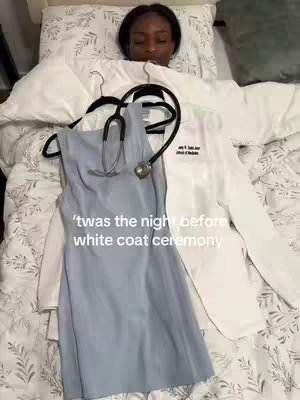 A young woman lies in bed, with the top of her head and face visible. Beside her on the bed is a light blue dress, a white lab coat with writing on the left breast pocket, and a stethoscope draped over the dress. The bedding is white with a subtle floral pattern.