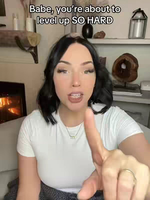 A medium shot features a fair-skinned woman with long dark hair wearing a white t-shirt, pointing her right index finger towards the viewer. Text overlayed on top reads, 'Babe, you're about to level up SO HARD'. The background includes a fireplace with a fire burning, a shelf with decorative items, and a couch