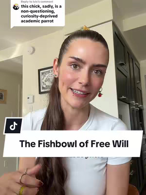 A woman with long brown hair and wearing a white t-shirt smiles while looking directly at the camera. A overlay at the bottom says "The Fishbowl of Free Will". In the background there is an interior setting with framed artwork.