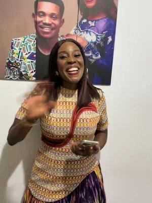 A woman with dark hair smiles and waves at the camera. She is wearing a patterned, colorful top and a long skirt with mixed colors. Behind her are poster portraits of a man and a woman, and the setting appears to be indoors.