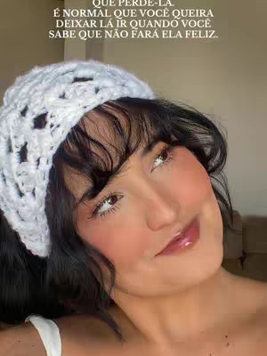 A close-up shot featuring a light-skinned woman with dark hair and a white, crocheted headband. She has a soft, rosy blush on her cheeks and is looking slightly upwards with a subtle smile.  The background is blurred, suggesting a neutral-toned interior.